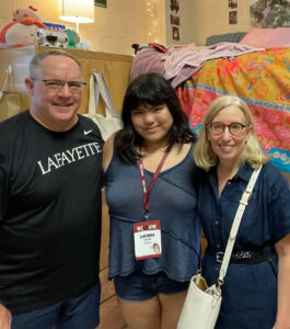 Kevin and Bethany Durkin with their daughter, Lucy ‘27, at Move-In. They are standing in Lucy's dorm. There are string lights, and photos in the background.