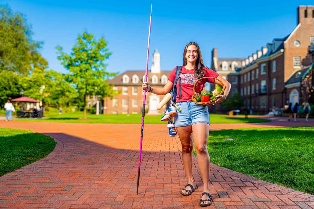 Rylee Bordwick '25 is holding a javelin in one hand and a basket of produce in the other. She is wearing shorts and has a backpack with cleats on her right shoulder.