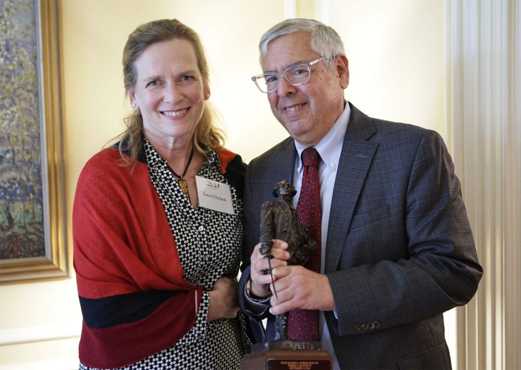 Laura Chylack and Robert J. Bliwise '76 are smiling at the camera. Robert is holding the mini statue of the Marquis de Lafayette. 